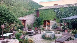 Een terras buiten met stenen tafels en stoelen, omgeven door weelderige begroeiing en een stenen muur.