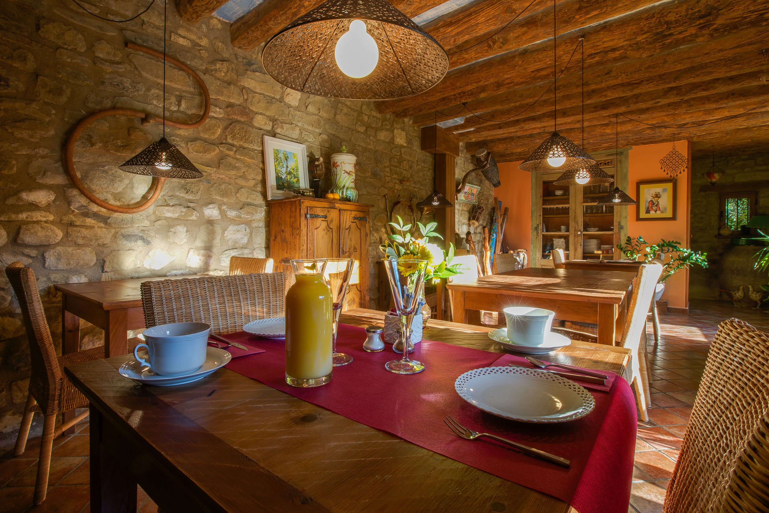 A rustic dining area with a wooden table set for two, surrounded by wicker chairs and a stone wall.