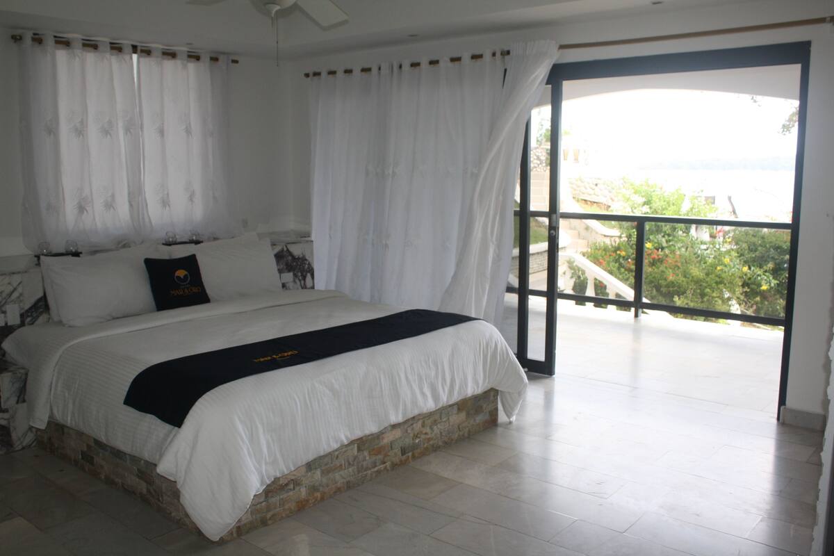 A bedroom with a large bed, white curtains, and a view of greenery through a glass door.
