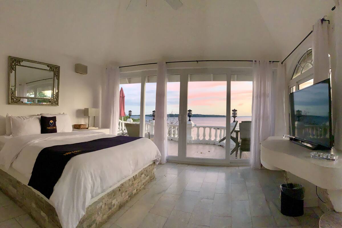 A hotel room with a bed, a television, a balcony with a view of the sea, and a large mirror on the wall.