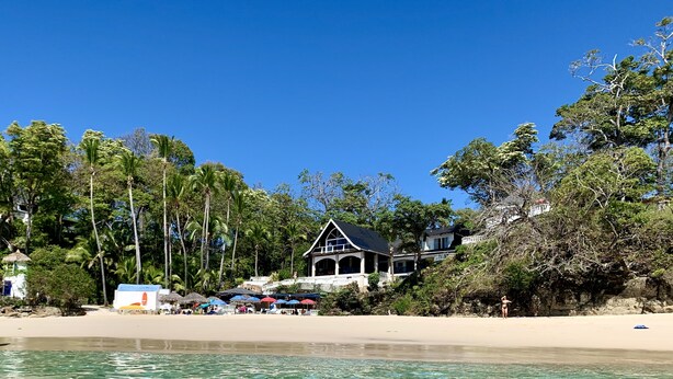 Une propriété située en bord de mer, avec une eau cristalline, des palmiers et une grande maison dotée d’un balcon.