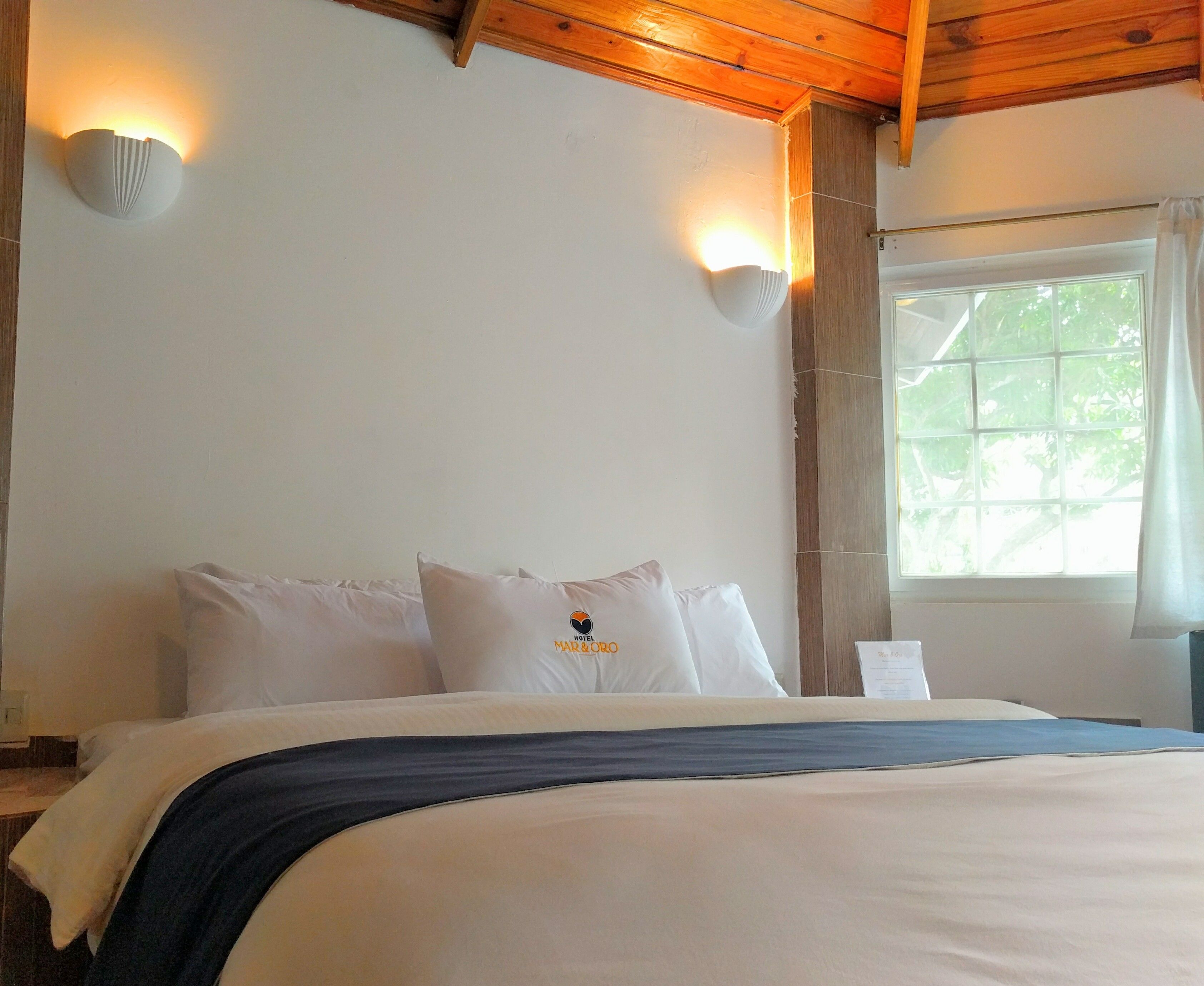 Una habitación de hotel con cama, dos lámparas fijadas a la pared, una ventana con cortinas y techo de madera.