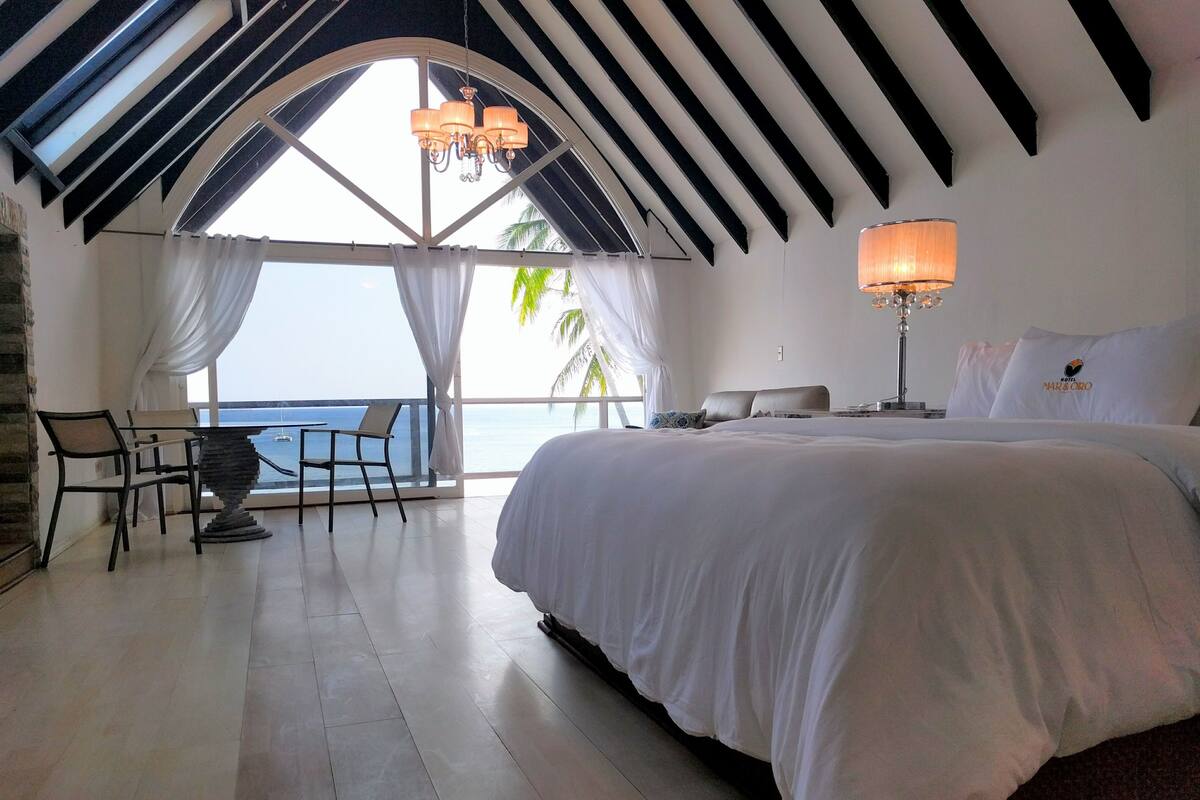 A hotel room with a large bed, a desk with a chair, a lamp, and a view of the sea.