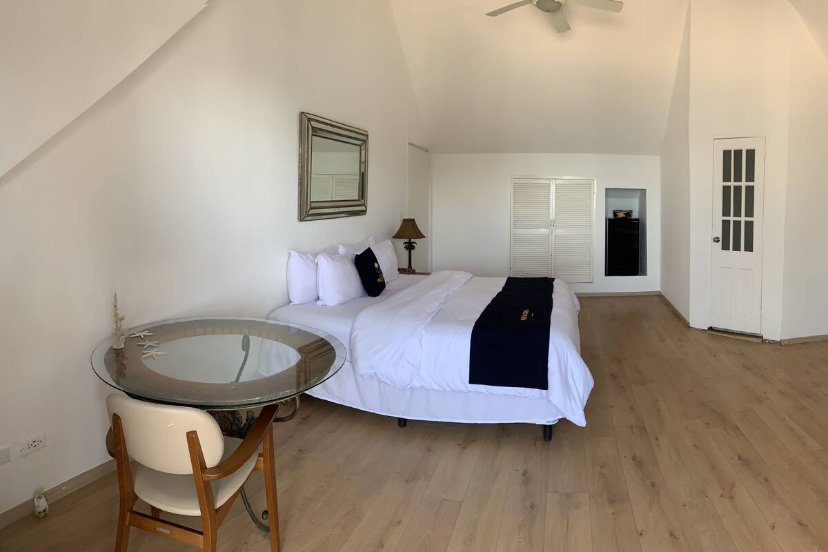 A bedroom with a bed, a glass table, a chair, a ceiling fan, a wardrobe, and a TV.