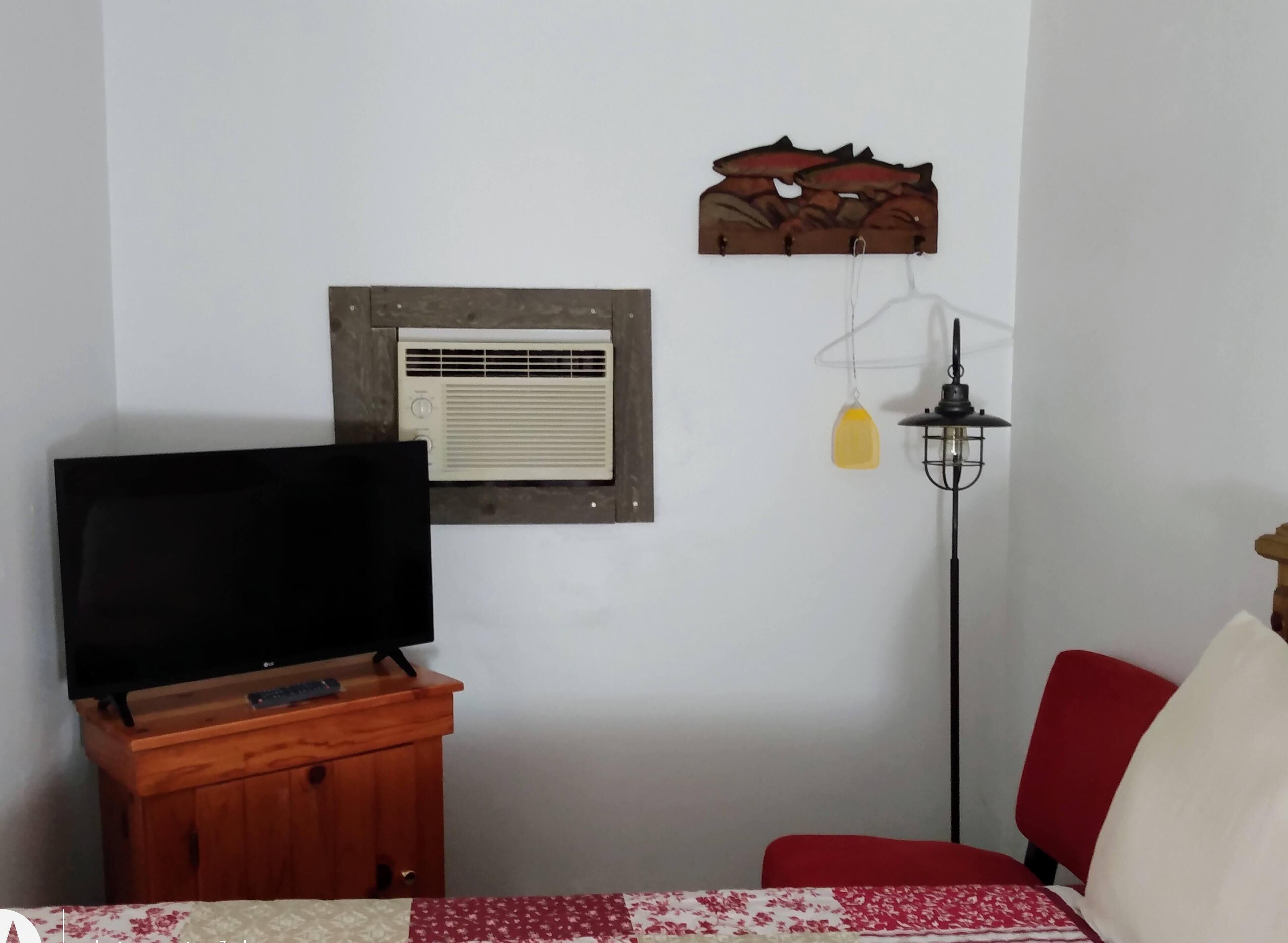 Un dormitorio con televisor sobre un mueble de madera, aire acondicionado de pared, lámpara colgante y una silla roja.