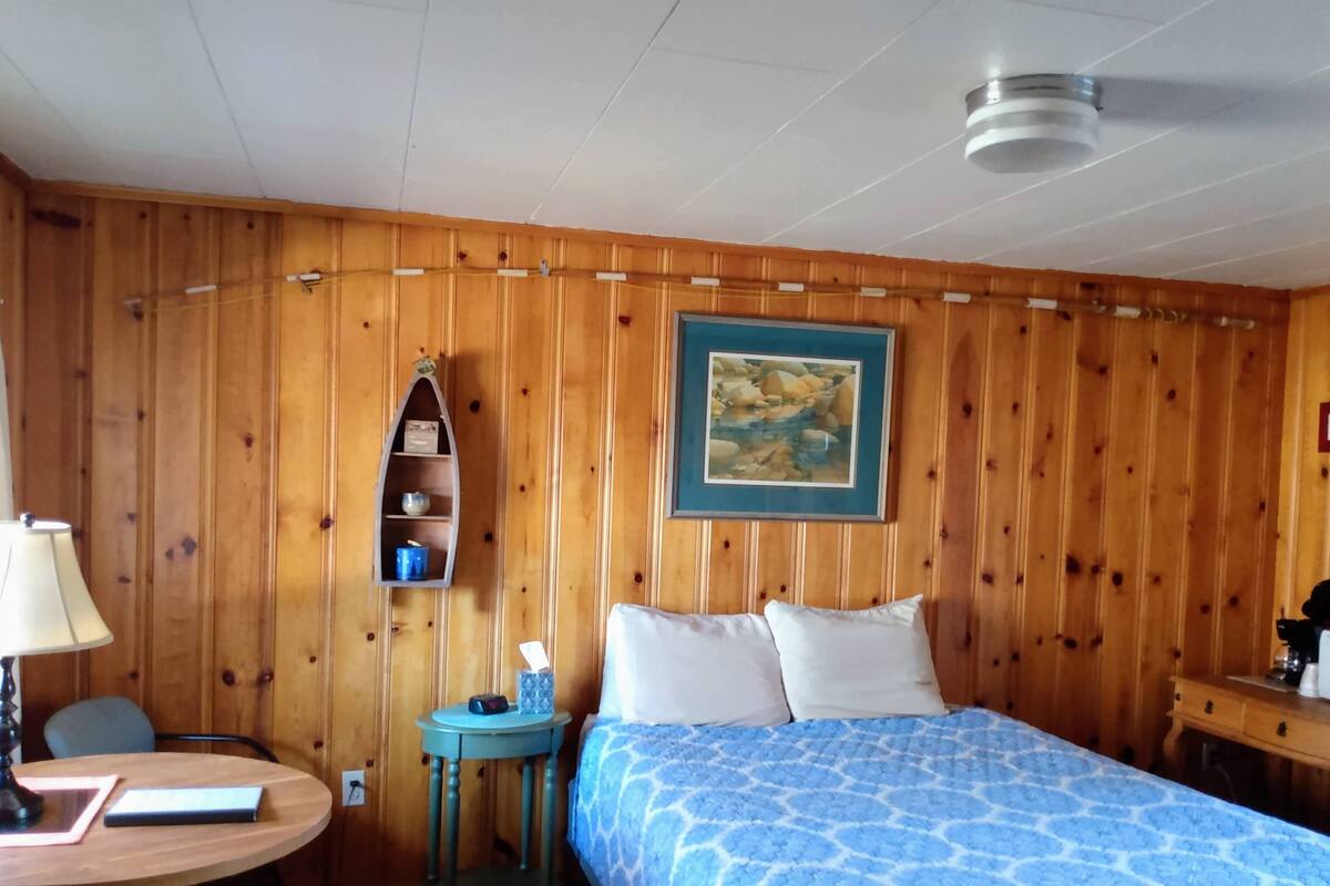 A wooden-paneled room with a bed, a small table with a lamp, a shelf with items, and a painting on the wall.