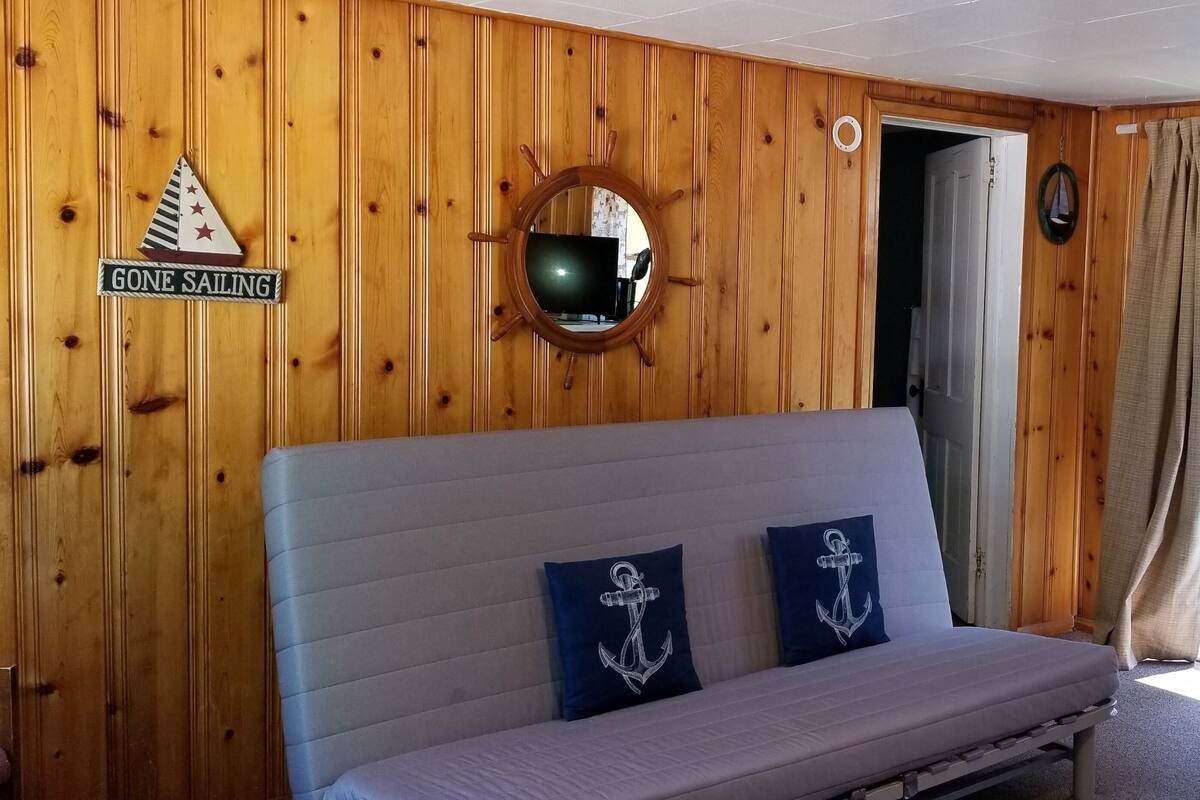 A cabin with a wooden interior, a sofa with anchor pillows, a round mirror, and a sign that reads "GONE SAILING."