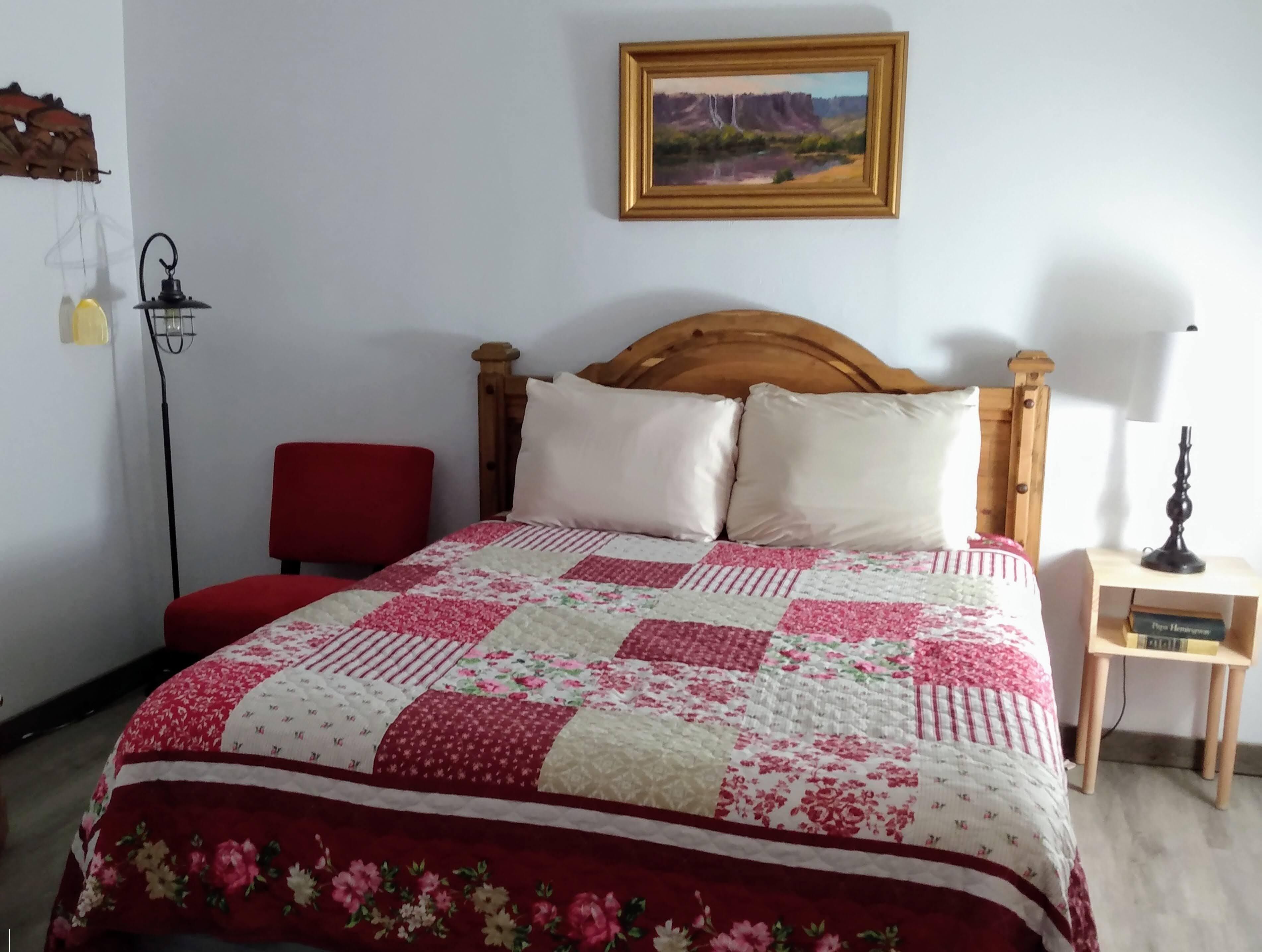 A bedroom with a wooden headboard, a quilted bedspread, a red chair, a bedside table with a lamp, and a framed painting on the wall.