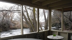 A room with a large window overlooking a snowy outdoor area with trees and a river. There is a round table and two chairs. - Billingsley Creek (Hagerman)
