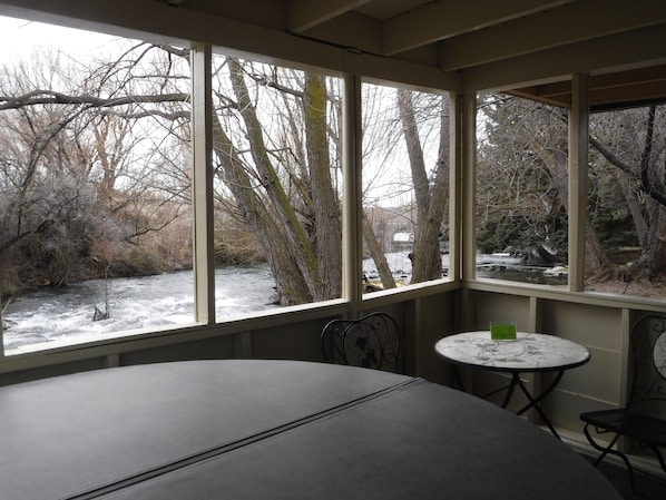 A room with a large window overlooking a snowy outdoor area with trees and a river. There is a round table and two chairs.