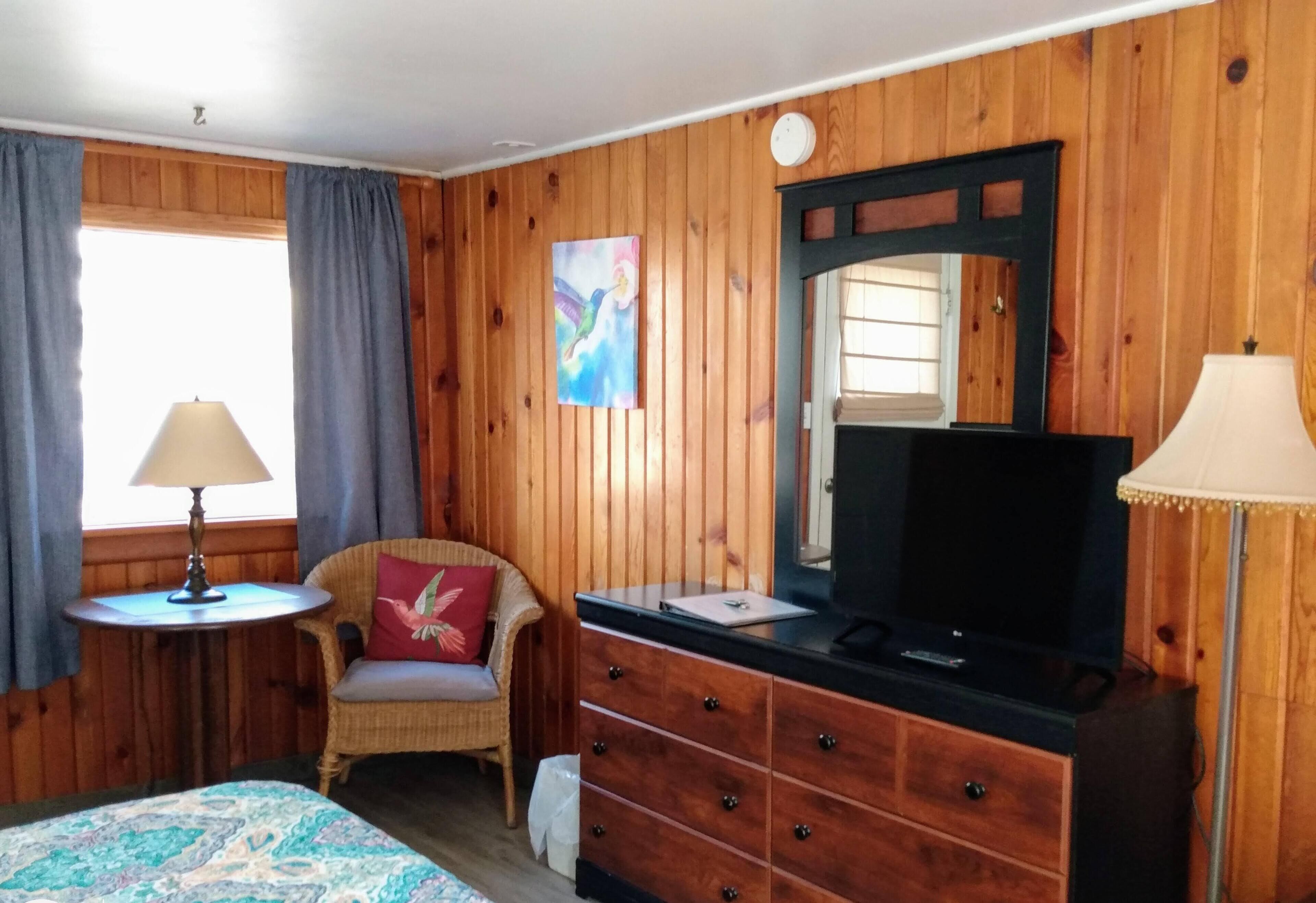 A room with wooden paneling, a bed with a patterned bedspread, a chair with a red pillow, a dresser with a mirror, a television, and two lamps.