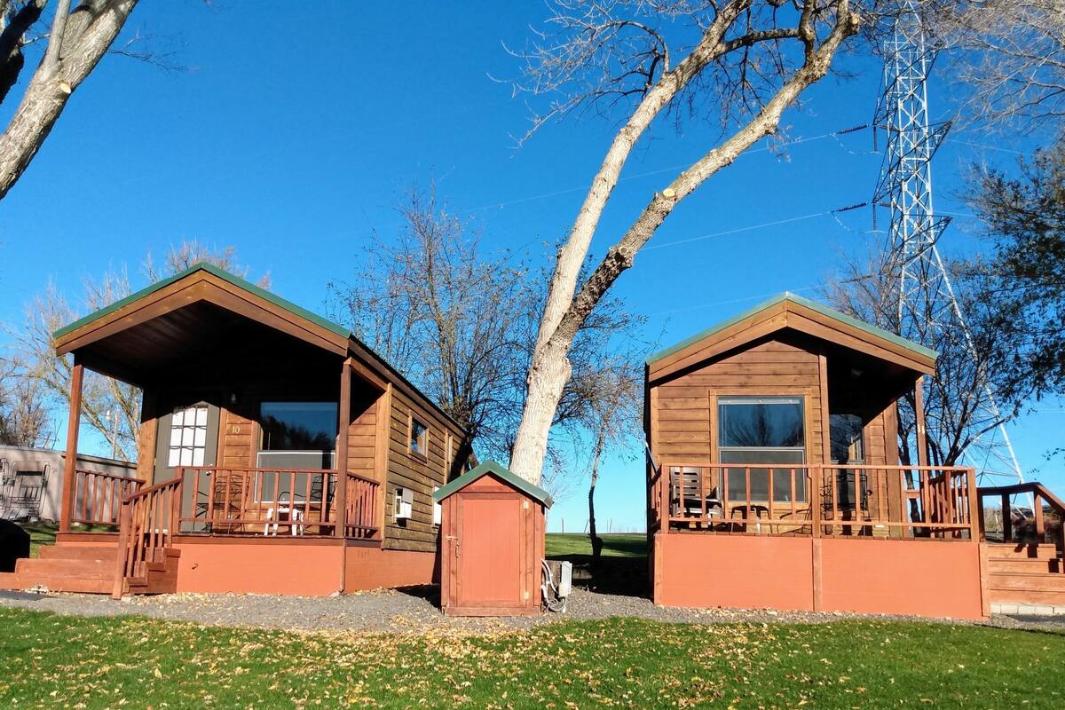Two wooden cabins with attached porches and a small shed, surrounded by trees and a clear sky.