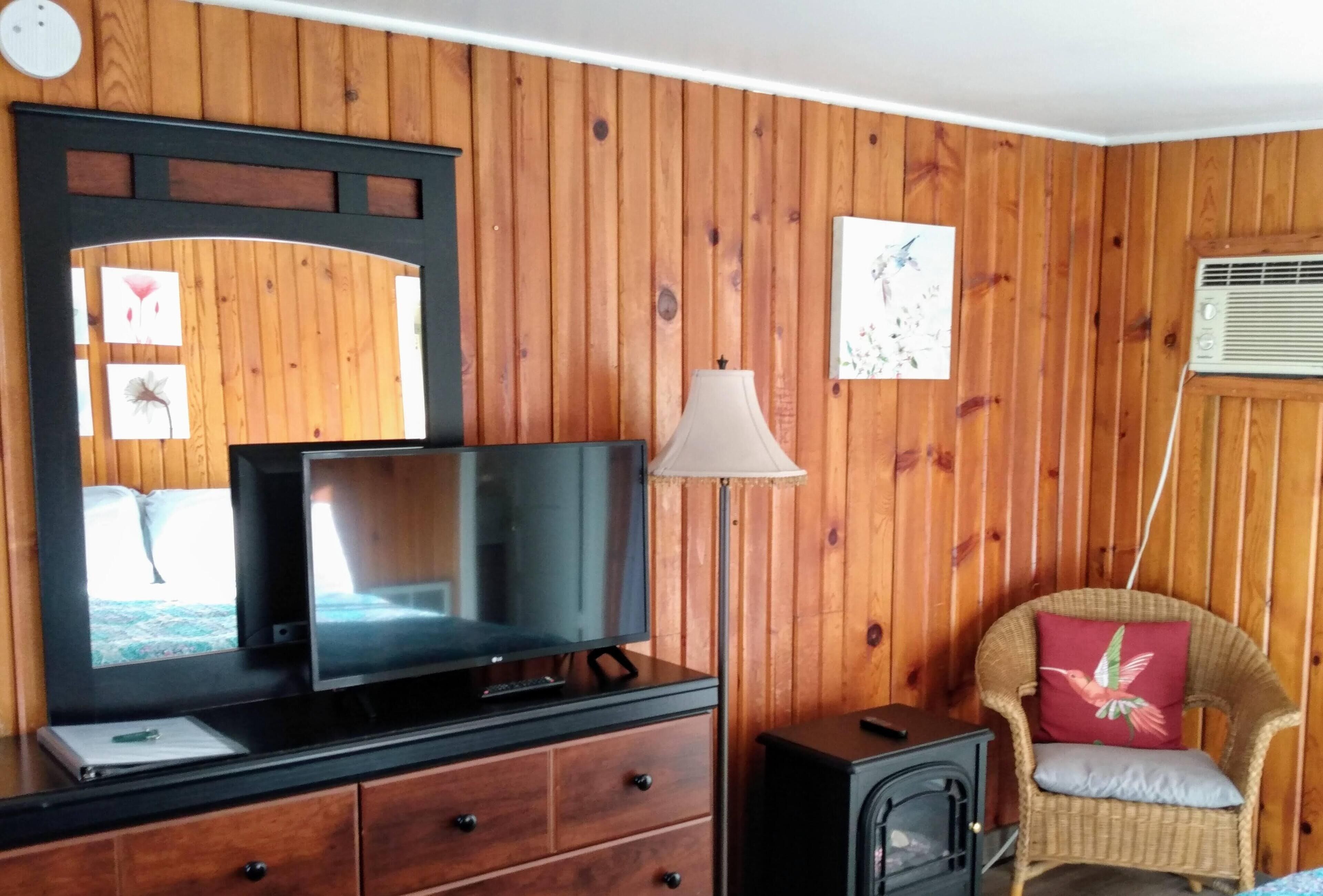 A room with wooden panel walls, a television on a wooden cabinet, a lamp, a wicker chair with a cushion, and a small heater.