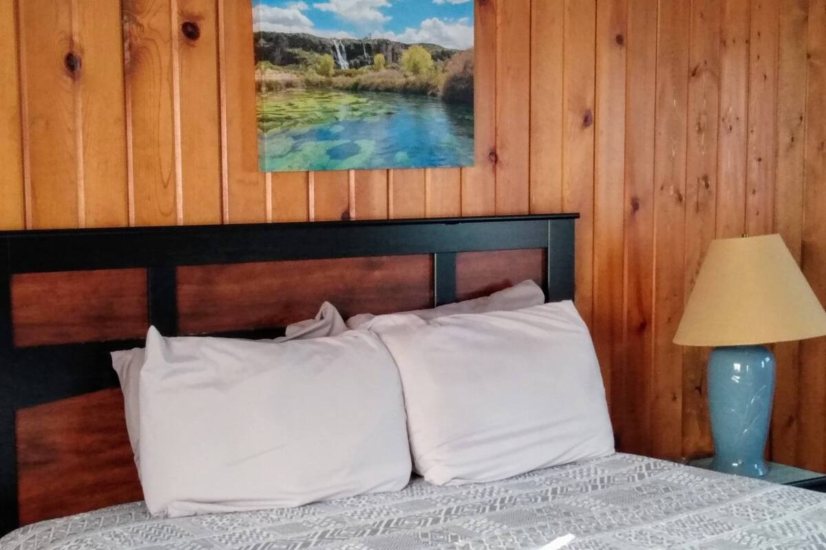 A bed with a patterned bedspread, two pillows, a wooden headboard, and a painting of a landscape above the bed.