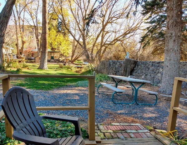 A wooden deck with a picnic table and chairs, surrounded by trees and a stone wall.