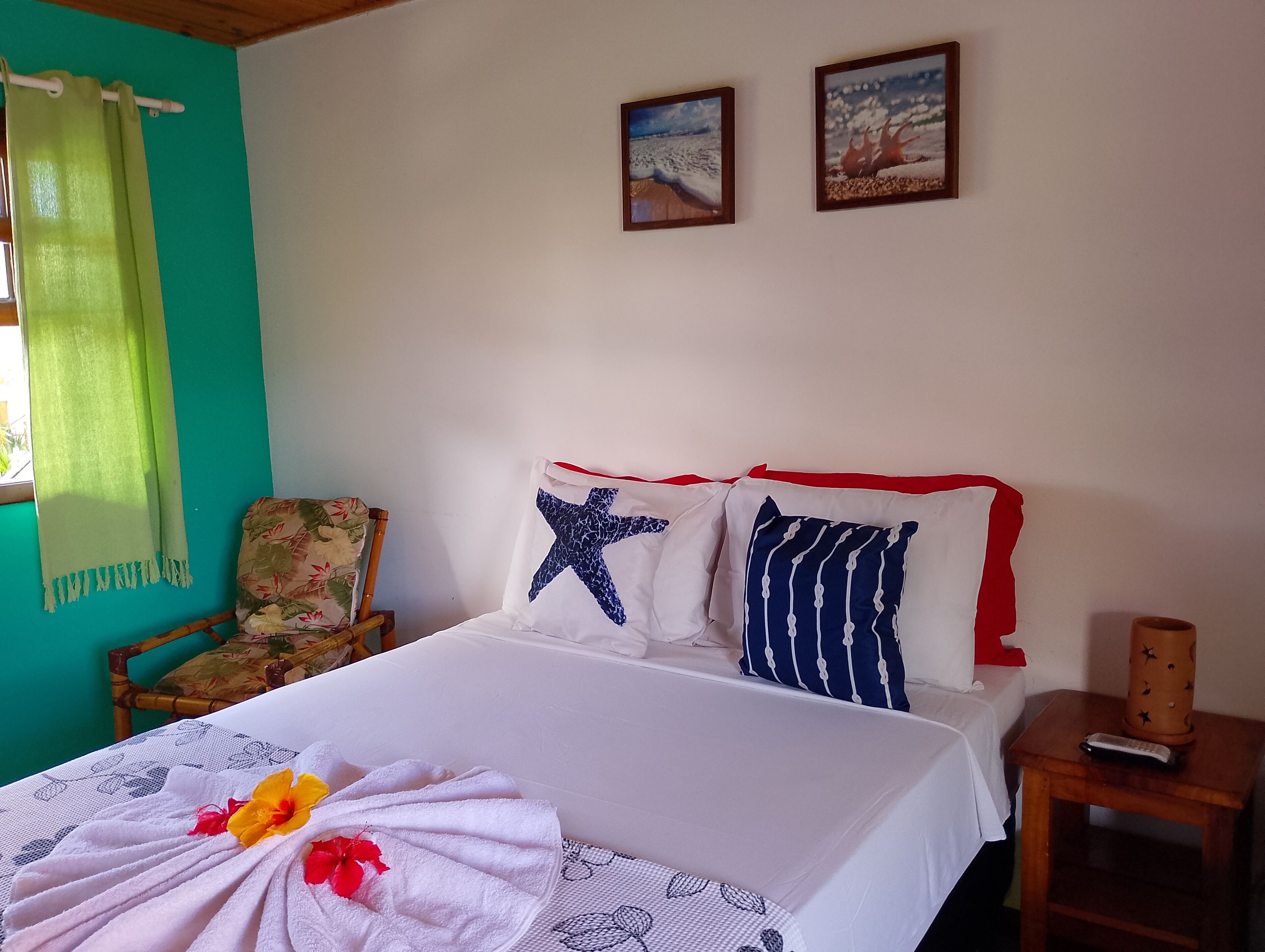 A neatly made bed with a floral towel and a starfish pillow, a wooden chair, and a small table with a remote control.