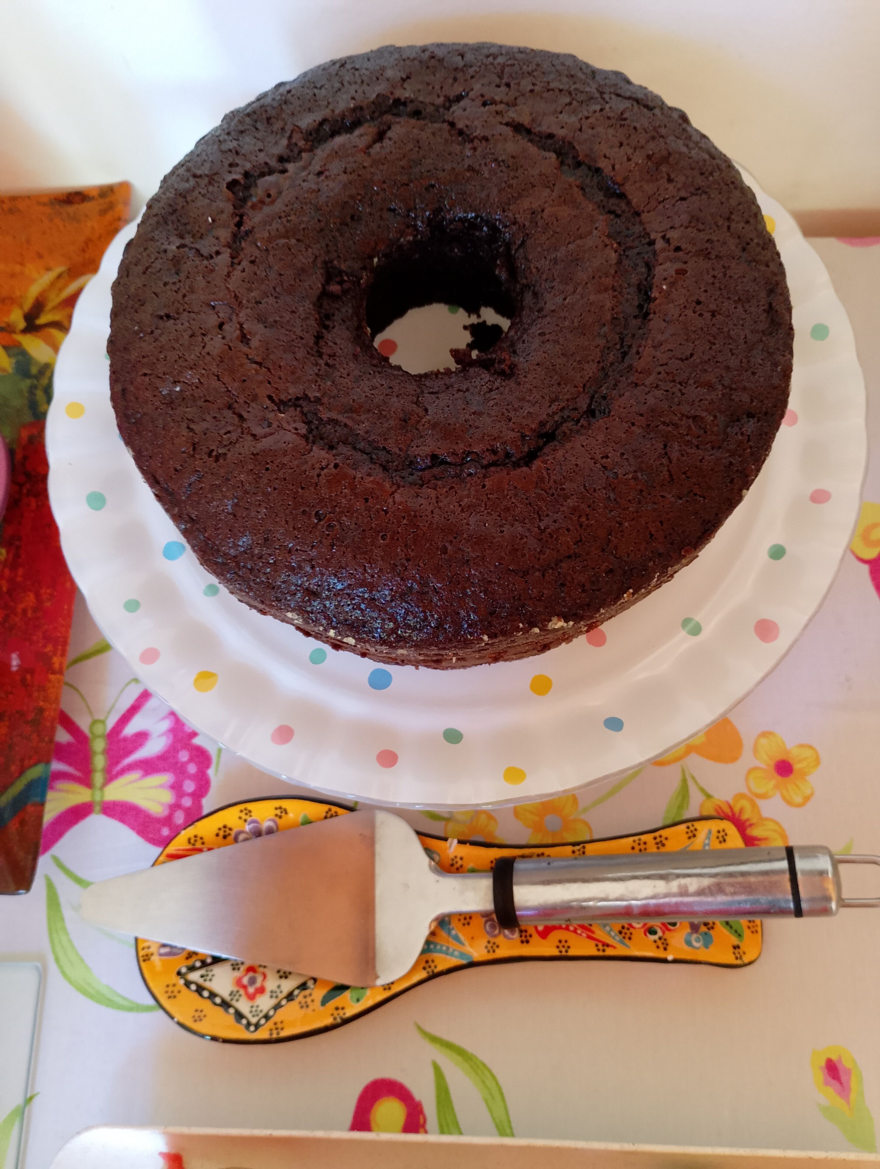A chocolate cake on a white plate with colorful polka dots, placed on a table with a floral tablecloth.