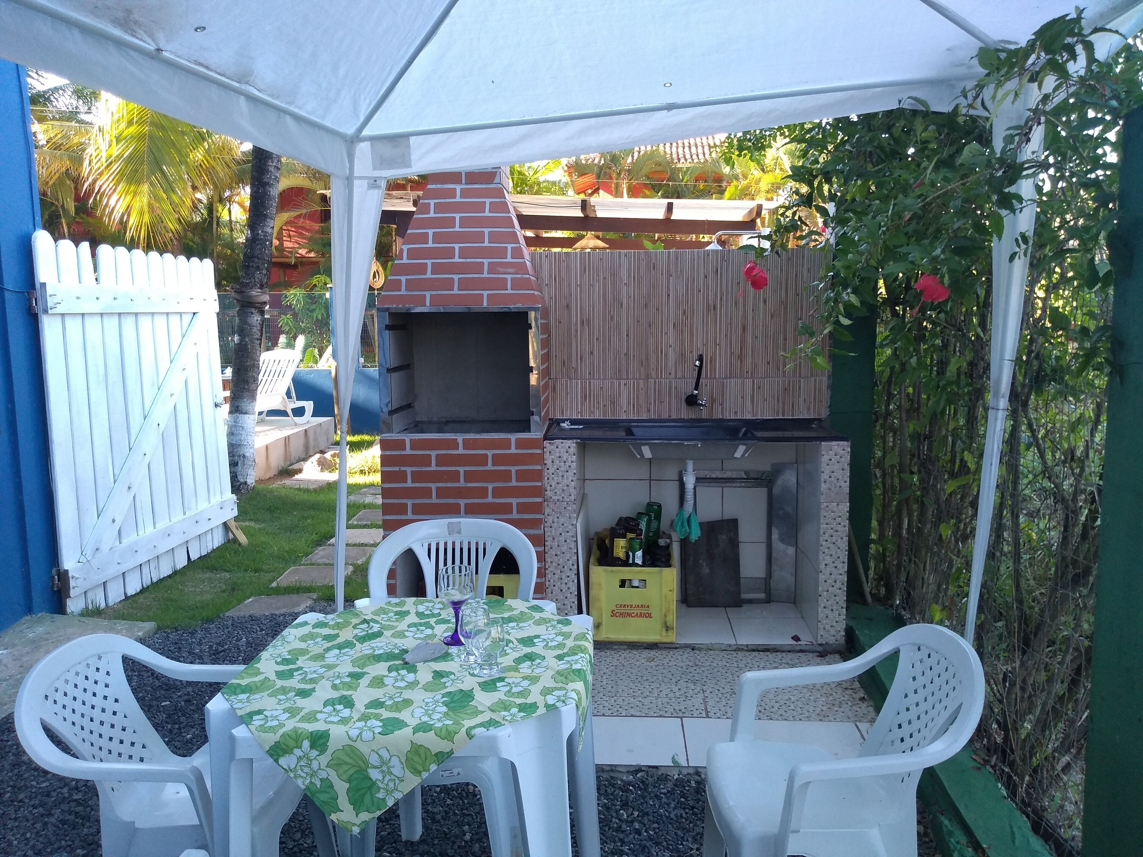 Eine Terrassenfläche mit einem gemauerten Grill, einem Tisch mit einer Blumen Tischdecke und weißen Plastikstühlen.
