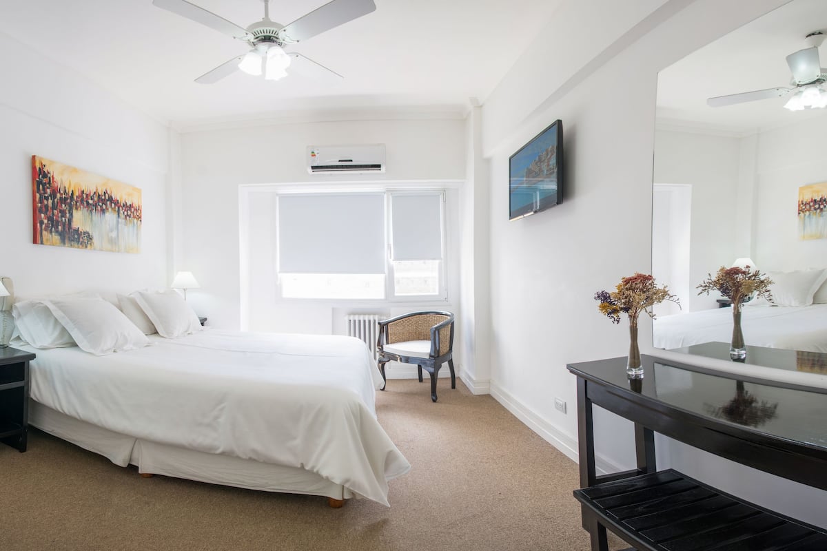 A hotel room with a bed, bedside tables, a chair, a desk, a mirror, and artwork on the walls.
