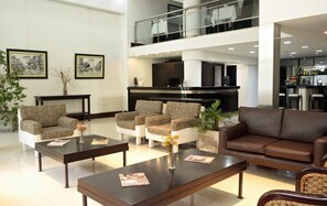 A modern hotel lobby with a reception desk, seating area with patterned sofas, a brown couch, and a bar area. - Hotel Corbel (Mar del Plata)