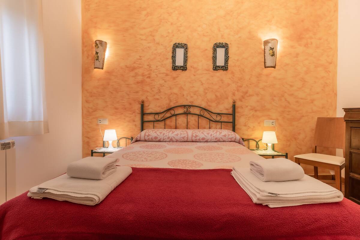 A neatly made bed with a red blanket, two bedside tables with lamps, and wall-mounted mirrors.