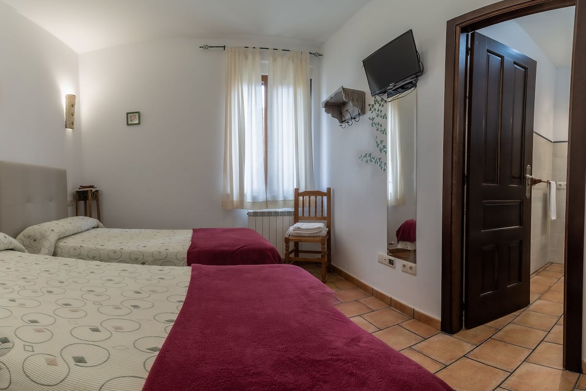 A bedroom with two beds, a nightstand with a lamp, a chair, a TV mounted on the wall, and a door.