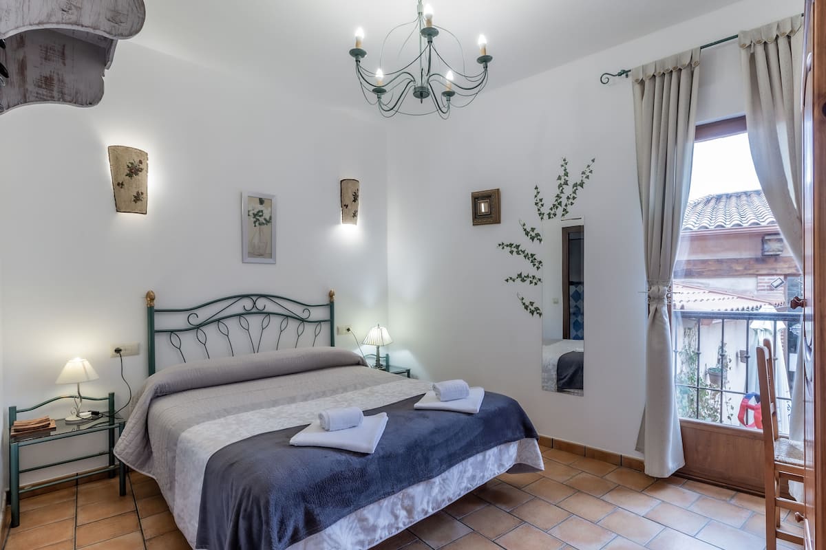 A bedroom with a bed, a nightstand, a lamp, a chandelier, and a window with curtains.