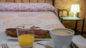 Una mesa de desayuno ordenada con un vaso de jugo de naranja, una taza de café y un plato de comida, sobre un edredón estampado y una mesita de noche con una lámpara.
