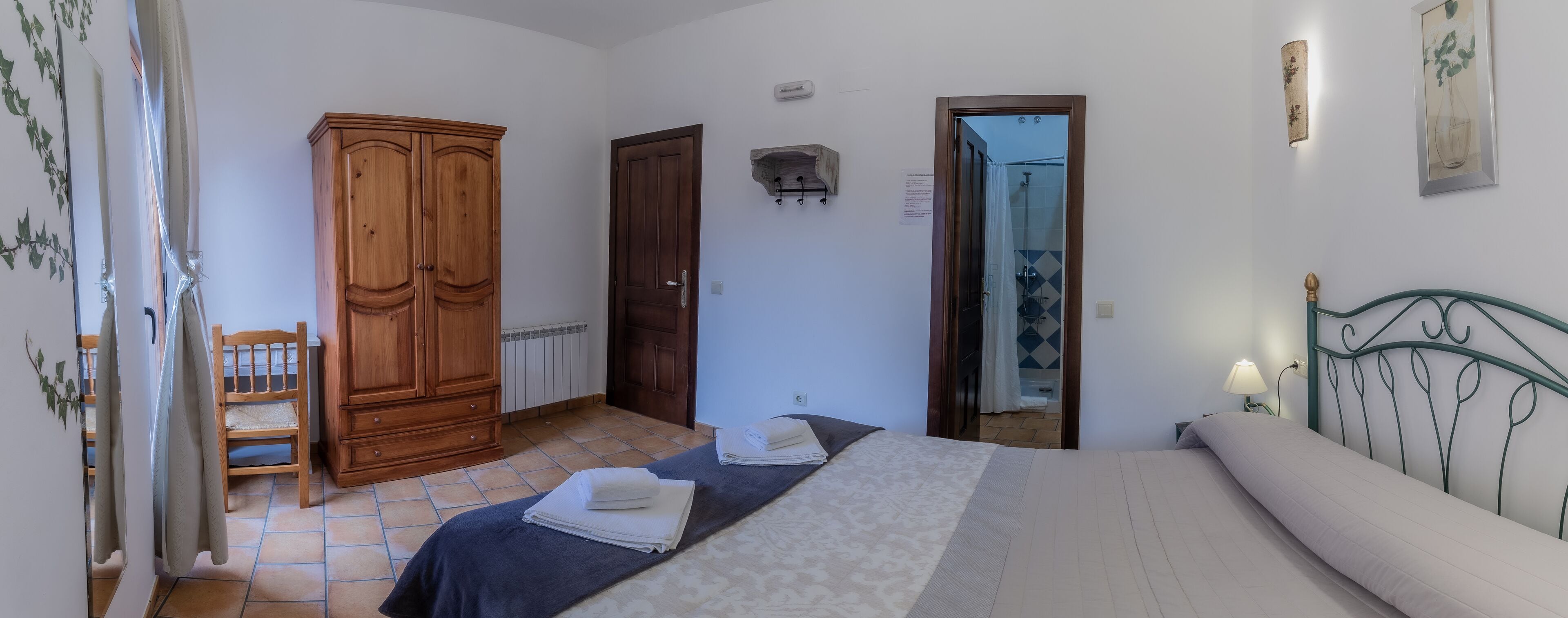 Un dormitorio con cama, armario, baño visible a través de una puerta abierta y una pequeña zona de estar con una silla.