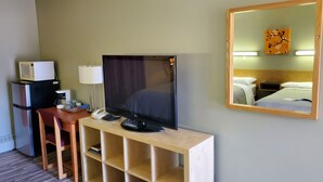 Et hotelværelse med fjernsyn på et træstel, mikrobølgeovn på et køleskab, et sengebord med lampe og en seng med sengegavl.