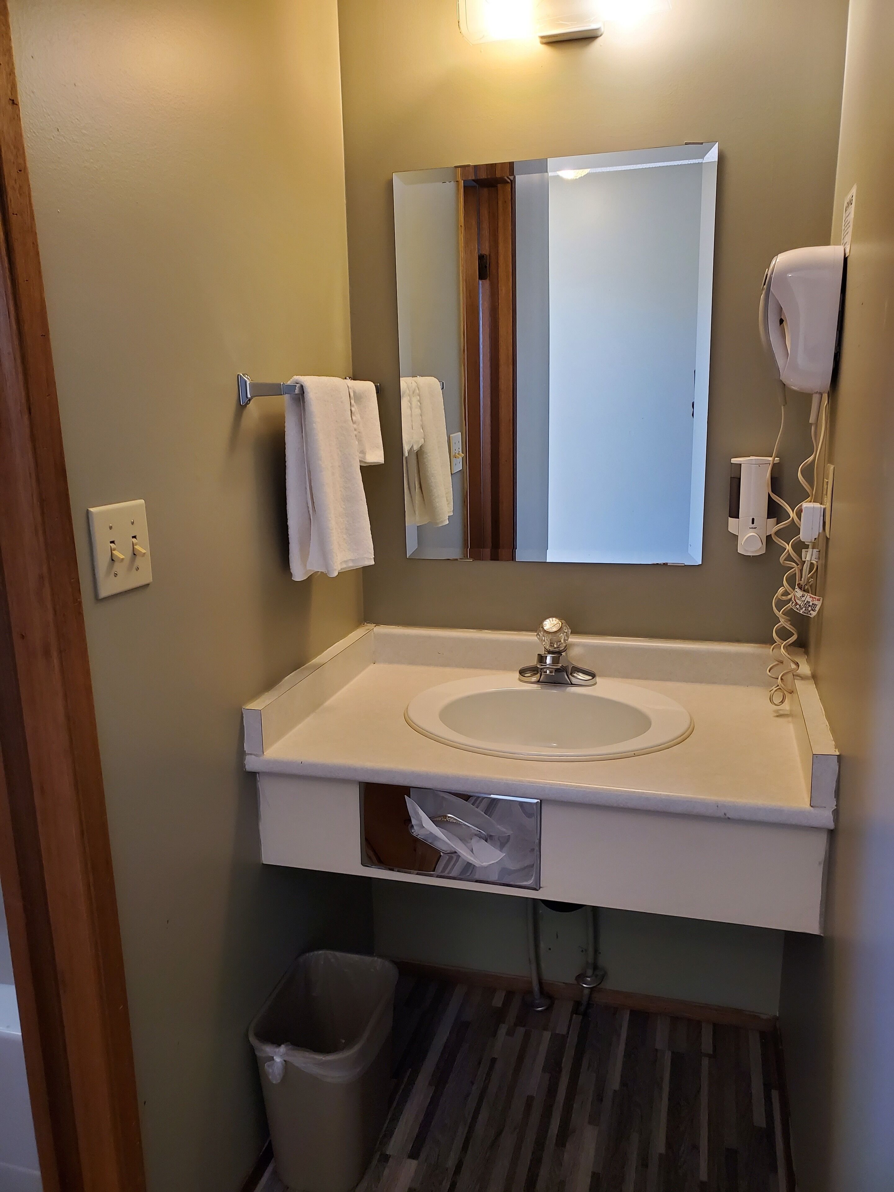 A bathroom with a white sink, mirror, and towel rack. There is a trash can and a hairdryer on the wall.