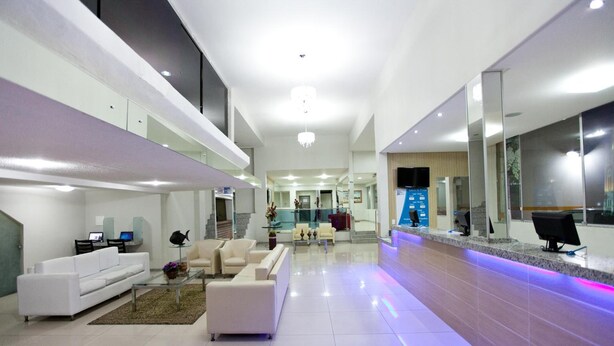 A spacious, modern hotel lobby with a high ceiling, a central seating area with sofas and armchairs, a reception desk, and a large TV screen. - JB Hotel (Petrolina)