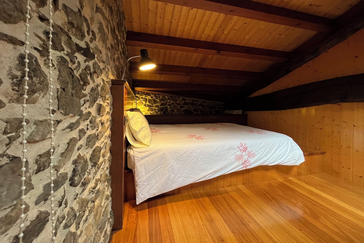 A bedroom with a wooden ceiling, stone walls, a bed with a white bedspread, and a bedside lamp.