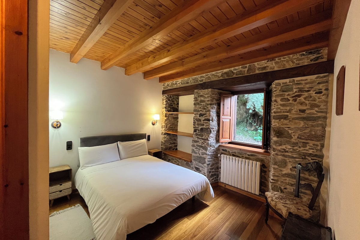 A bedroom with a wooden ceiling, stone walls, a bed, bedside lamps, and a window with a view of greenery.