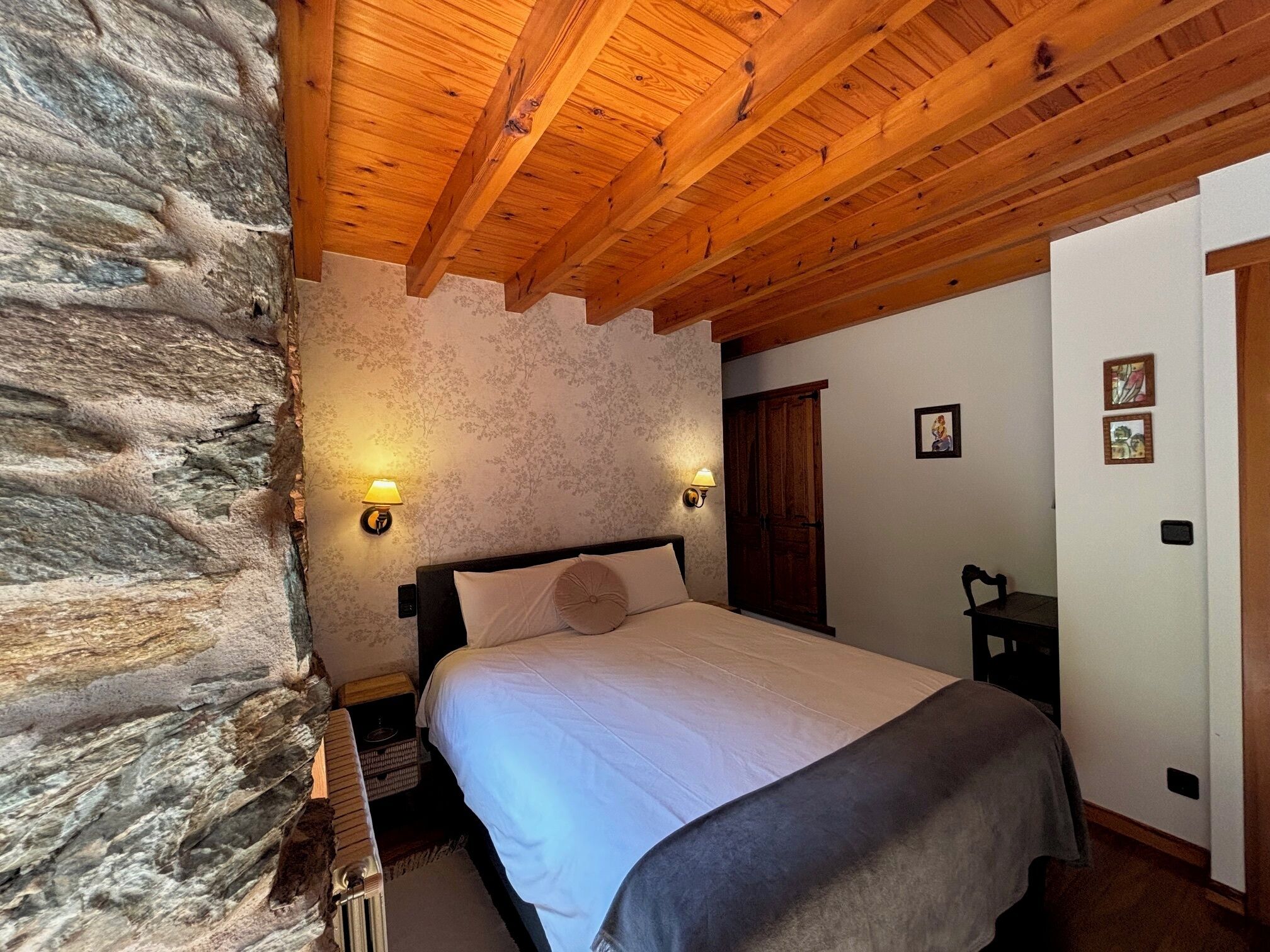 A bedroom with a stone wall, wooden ceiling beams, a bed with a white and gray bedspread, two wall-mounted lamps, and a wooden wardrobe.