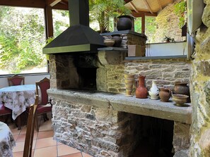 A stone-built outdoor kitchen with a chimney, surrounded by potted plants and rustic pottery. - Casa Rural O Vilar (Ortigueira)