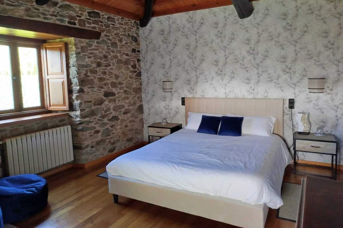 A bedroom with a stone wall, wooden ceiling beams, a bed with white bedding, two bedside tables, and a window with a view.