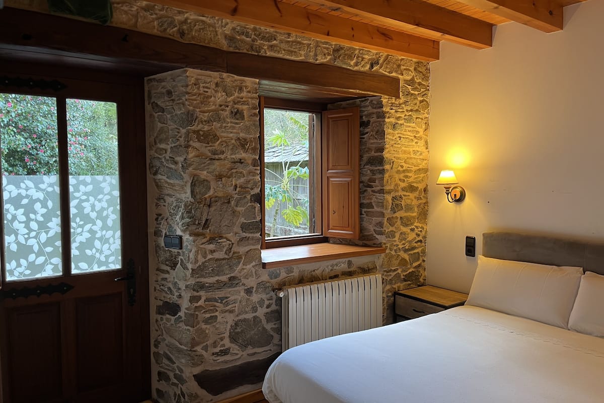 A bedroom with stone walls, a wooden ceiling, a window with a wooden frame, a bedside table, a lamp, and a bed.