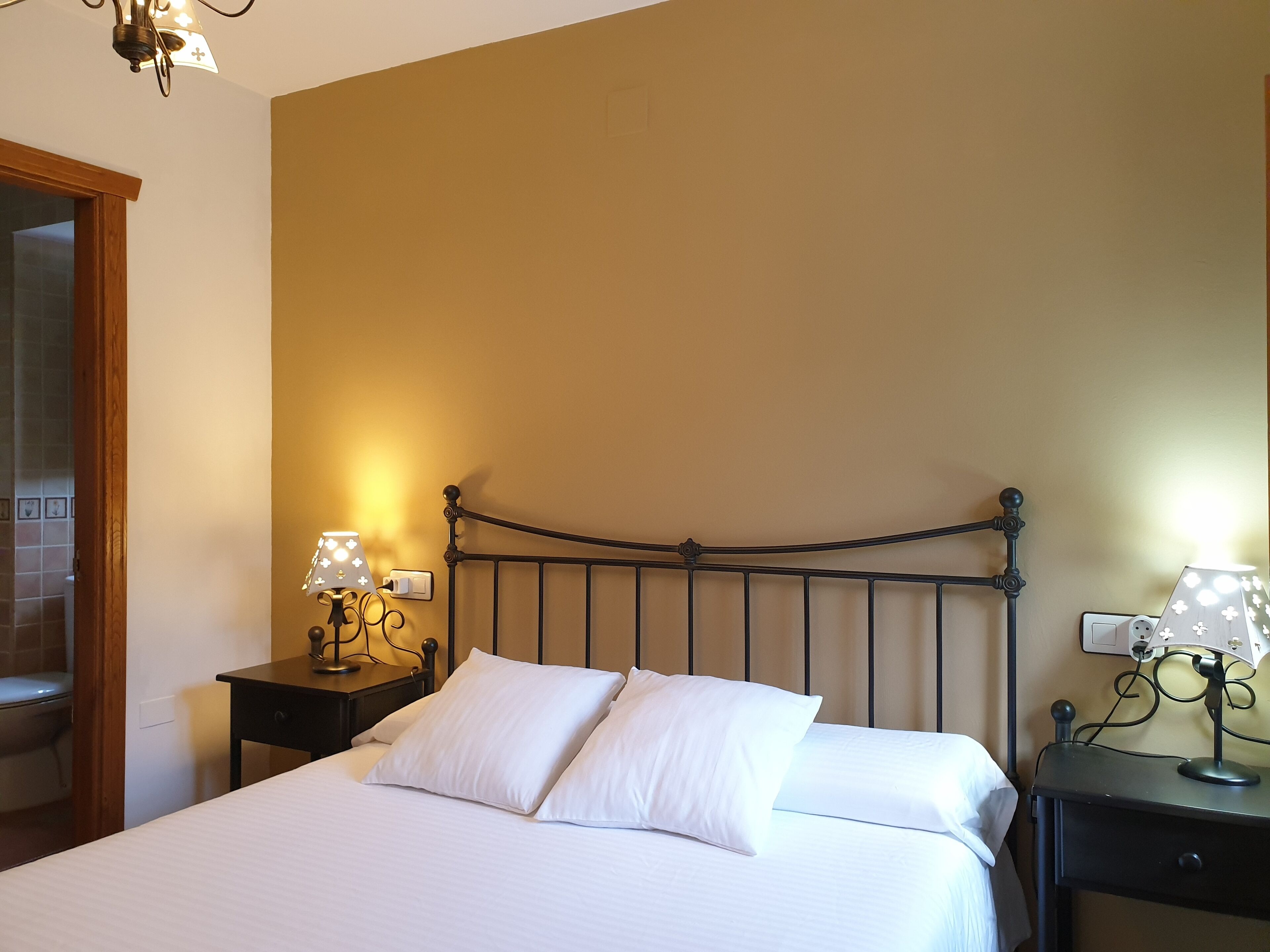 A neatly made bed with white linens in a room with beige walls and a metal headboard. There are two bedside tables with lamps, and a wooden door is visible to the left.