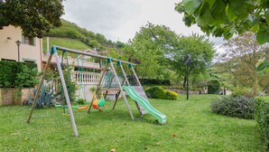 A playground with swings and a slide, surrounded by grass and trees. - La Pumarada de Limés (Cangas del Narcea)