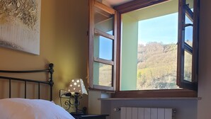 A neatly made bed with white linens, a colorful crocheted throw, a nightstand with a lamp, and a window with a view of a forested landscape. - La Pumarada de Limés (Cangas del Narcea)