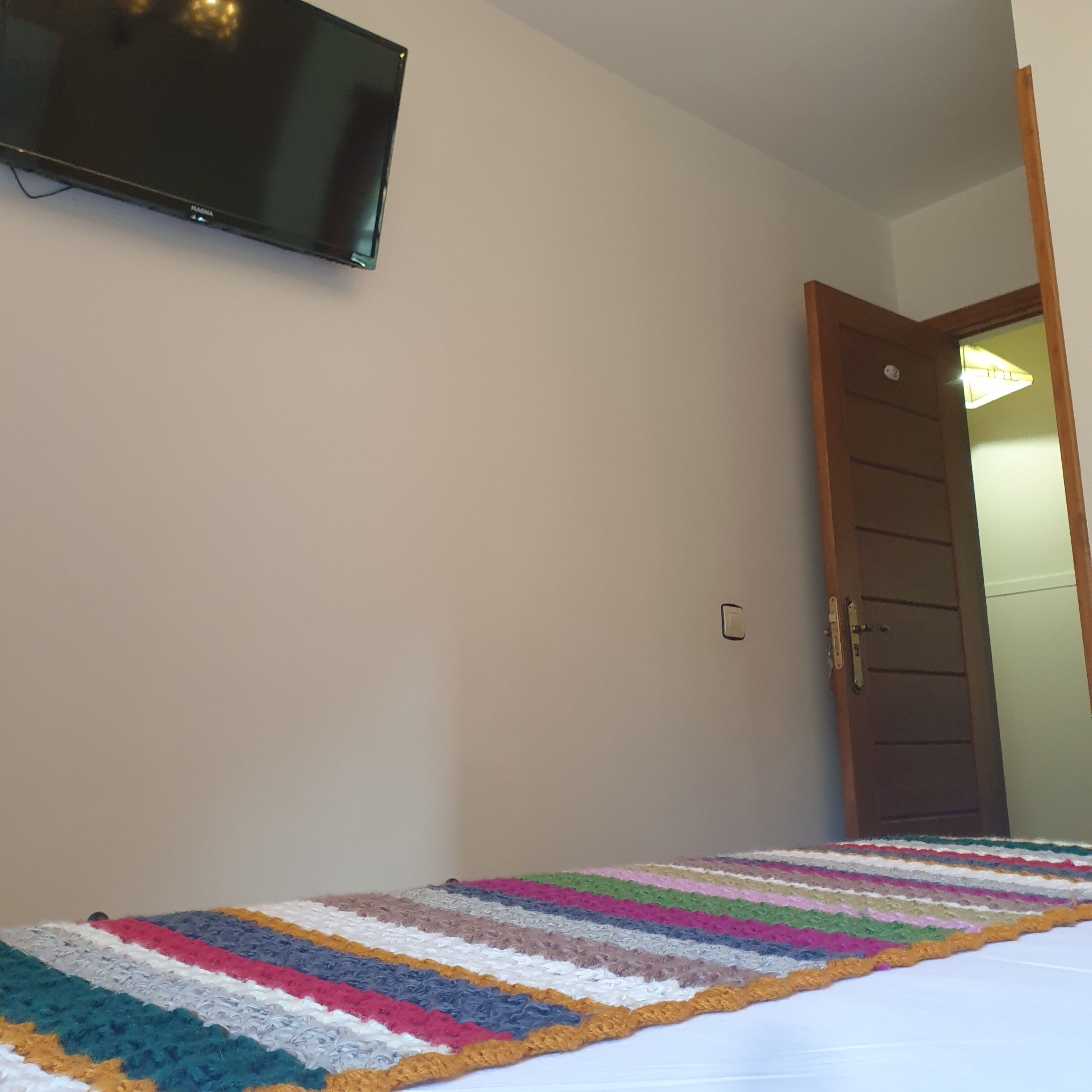 A room with a television mounted on the wall, a wooden door, and a colorful striped blanket on the floor.