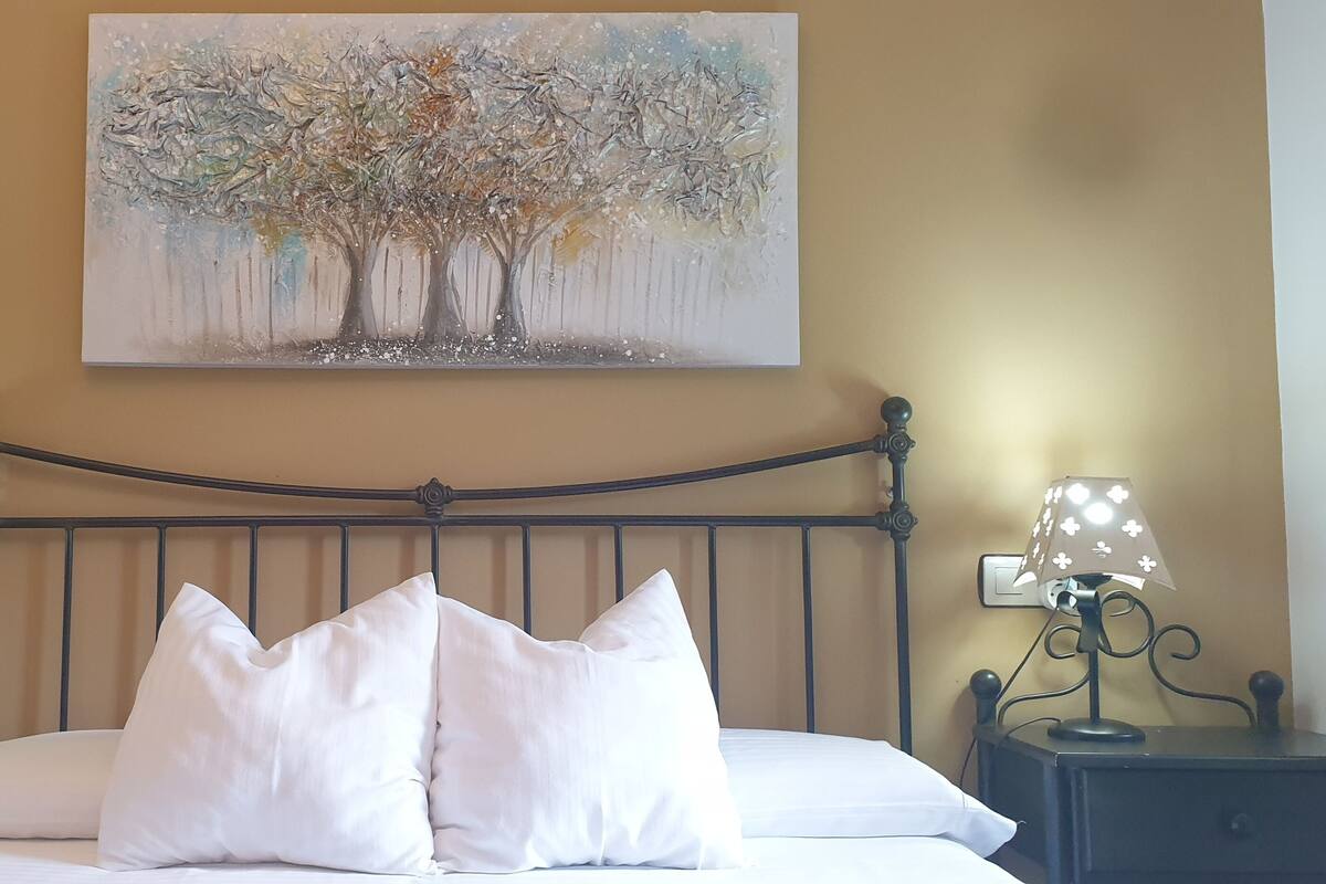 A neatly made bed with white linens and two pillows, flanked by two bedside tables with lamps and a painting of a tree on the wall above.