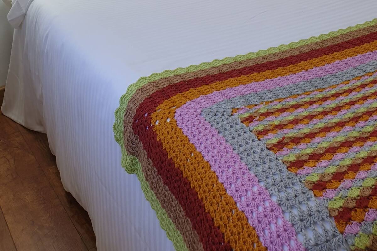 A neatly made bed with white linens and a colorful crocheted blanket, a white headboard, and a wall-mounted nightlight.