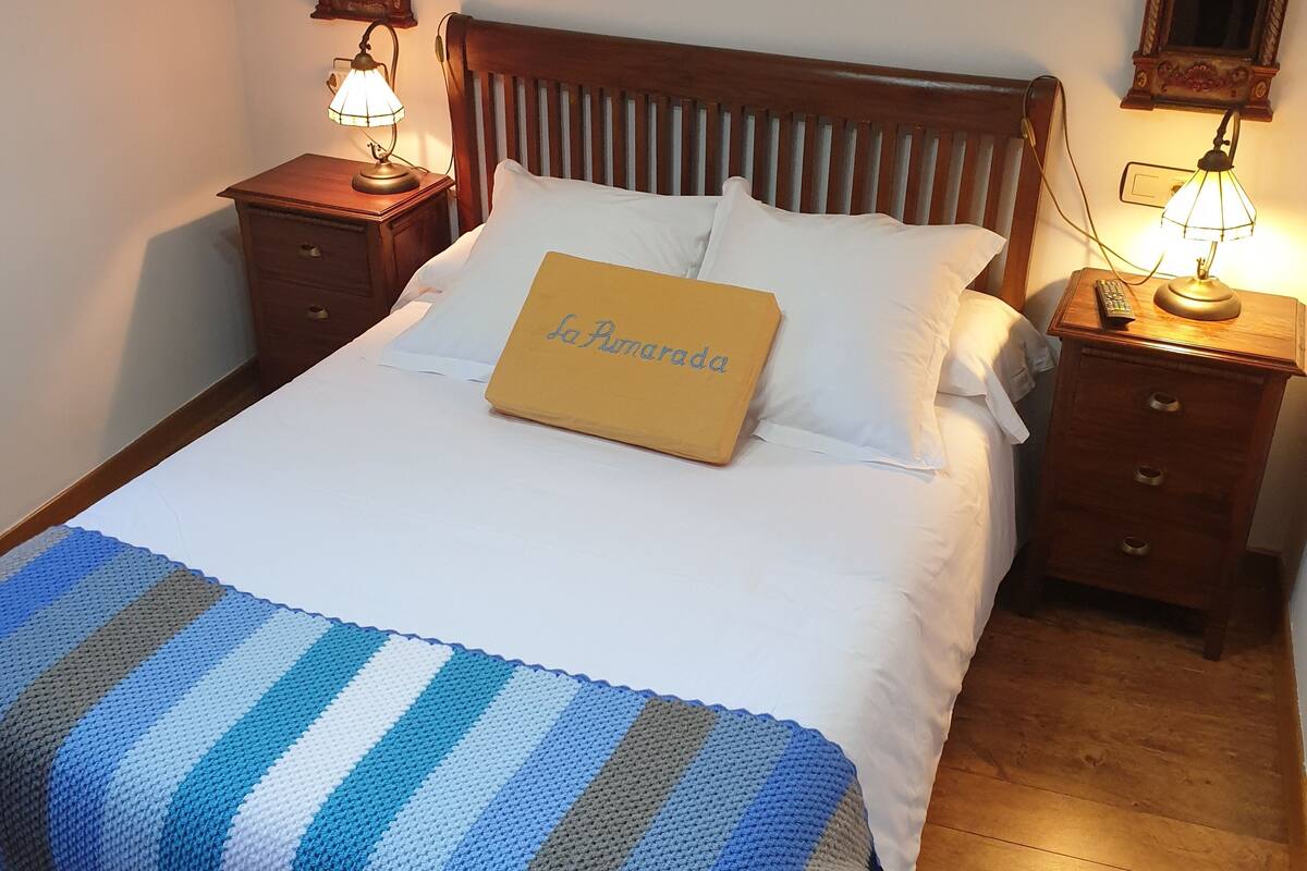 A neatly made bed with a striped blanket, two bedside lamps, and a pillow with "La Pomerada" text.