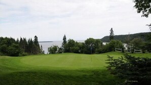 A golf course with a water body in the background, surrounded by trees and hills. - Hôtel Mingan (Sept-Iles)