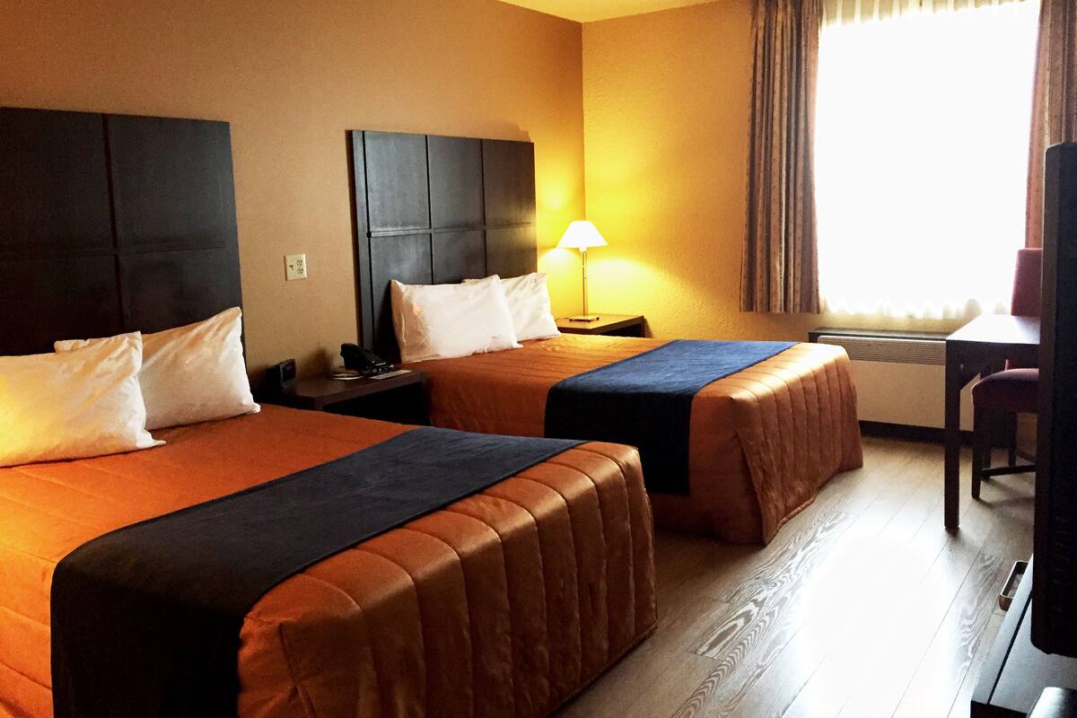 Habitación de hotel con dos camas, un escritorio, una silla, una lámpara y un ventanal con cortinas.