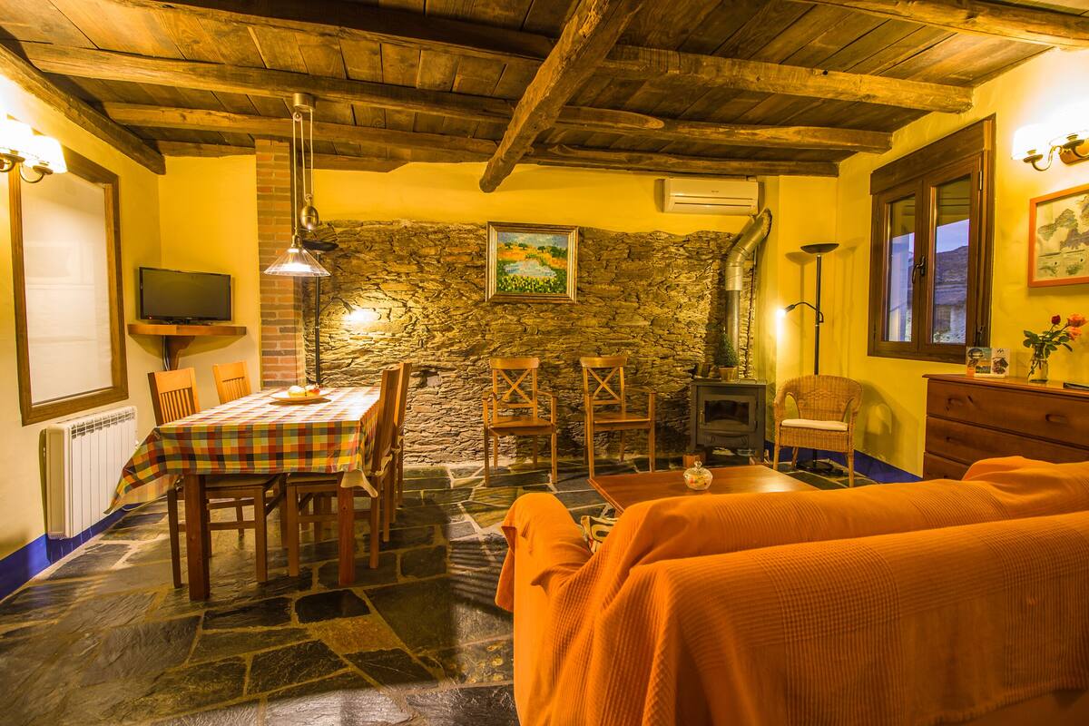 A cozy room with a stone wall, wooden ceiling beams, a fireplace, a table with chairs, and a bed with orange bedding.