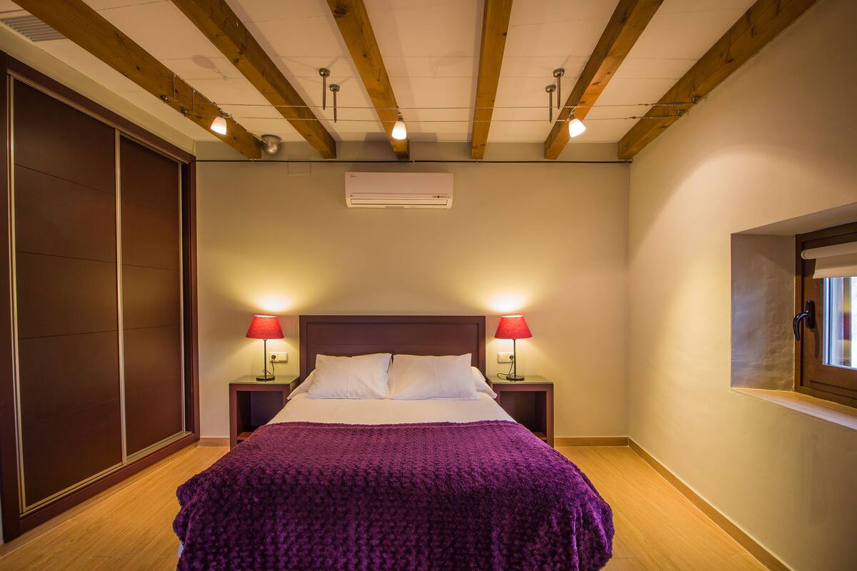 A bedroom with a wooden ceiling, a bed with a purple blanket, two bedside lamps, and a window on the right.
