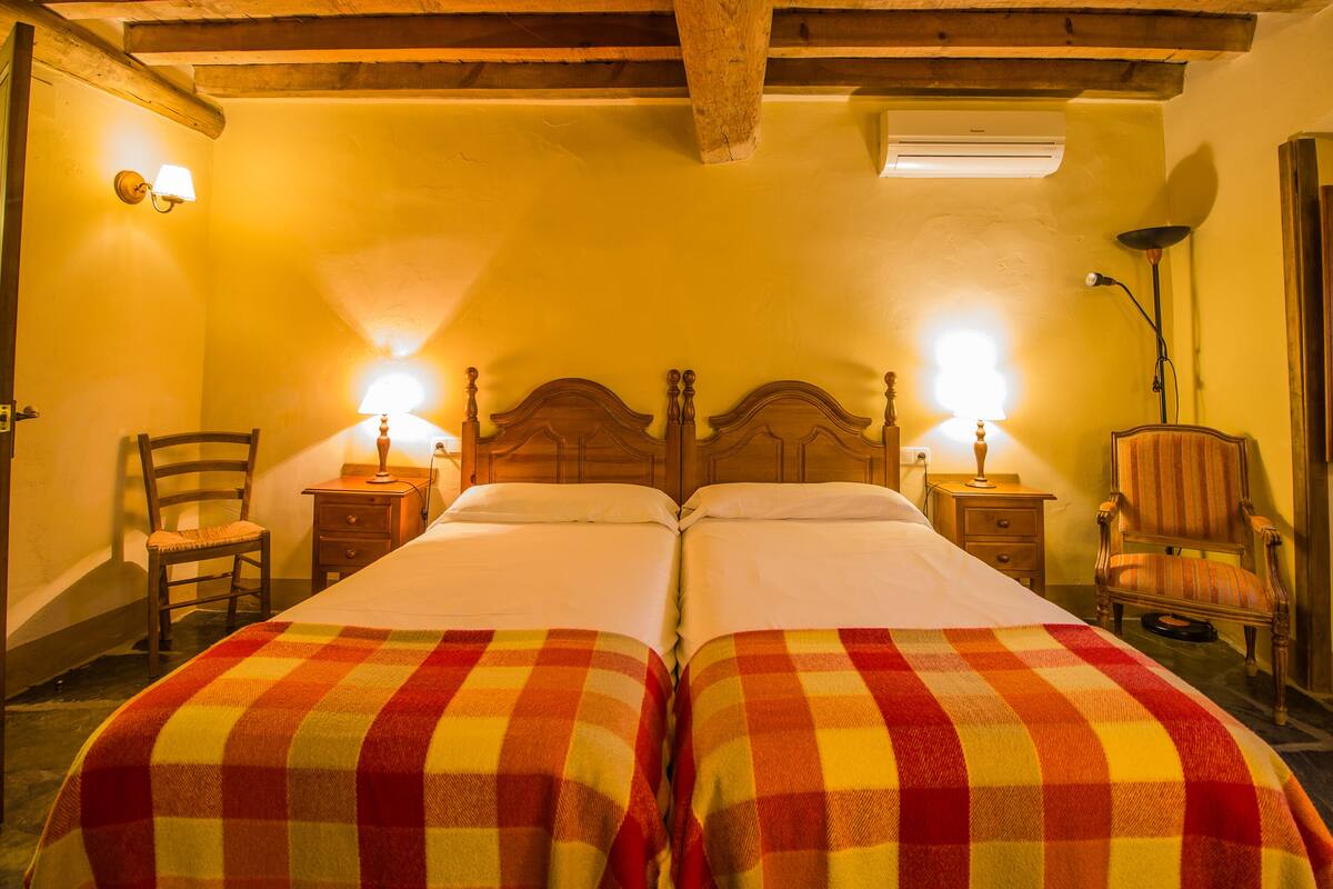 A room with two beds, each with a checkered blanket, wooden headboards, and bedside lamps.